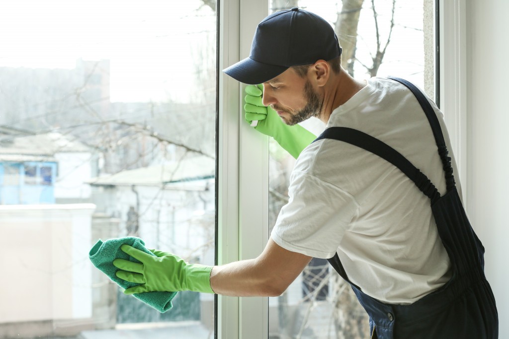 Man wearing black cap and green gloves cleaning home windows with cloth