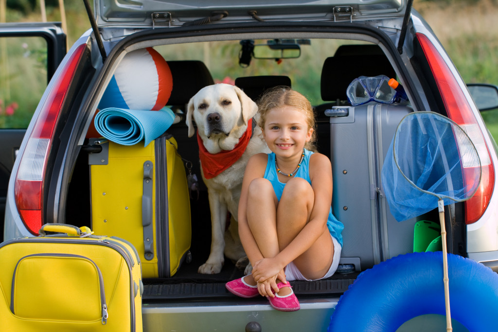 An automobile with an open trunk and several bags, as well as a dog and a young girl sitting inside.