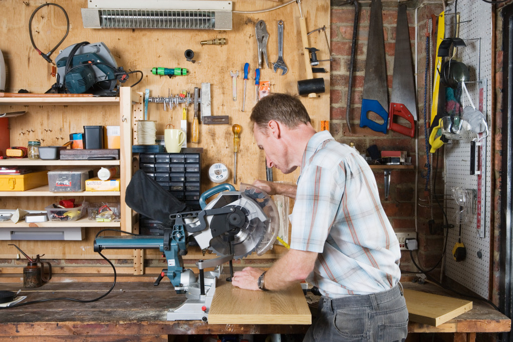A middle-aged man doing woodwork in the garage using a table saw with various tools hanging on the wall