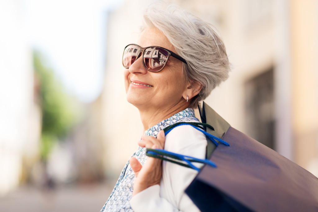 senior woman wearing sunglasses smiling while carrying shopping bags outdoors